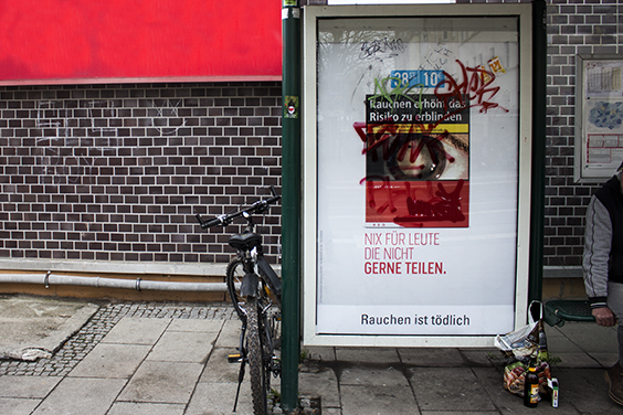 Haltestelle mit einem Werbekasten, in dem siche ein Werbeplakat für den Zigarettenhersteller Gauloises befindet. Der Markenname wurde mit Graffiti-Tags unkenntlich gemacht.