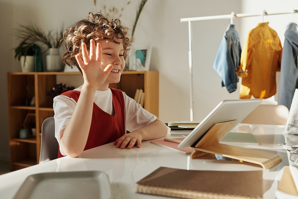 Ein Mädchen sitzt in ihrem Zimmer und nimmt an einer Online-Unterrichtsstunde teil. Sie hebt ihre Hand und lächelt. Im Hintergrund sehen wir eine Kleiderstange mit Jacken, ein Schrank mit Büchern und anderen Gegenständen bestückt.