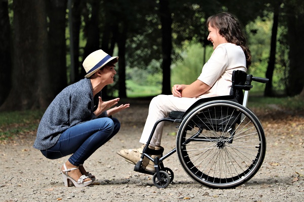 Zwei Frauen unterhalten sich auf einem Waldweg. Eine hockt, die andere sitzt im Rollstuhl. Beide lachen.
