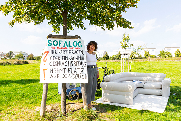 Stadträtin Dörte Jacobi posiert neben einem Baum, welcher das Poster des Sofalog trägt. Rechts neben ihr steht das Sofa des Sofalogs, dahinter ihr Fahrrad mit Anhänger.