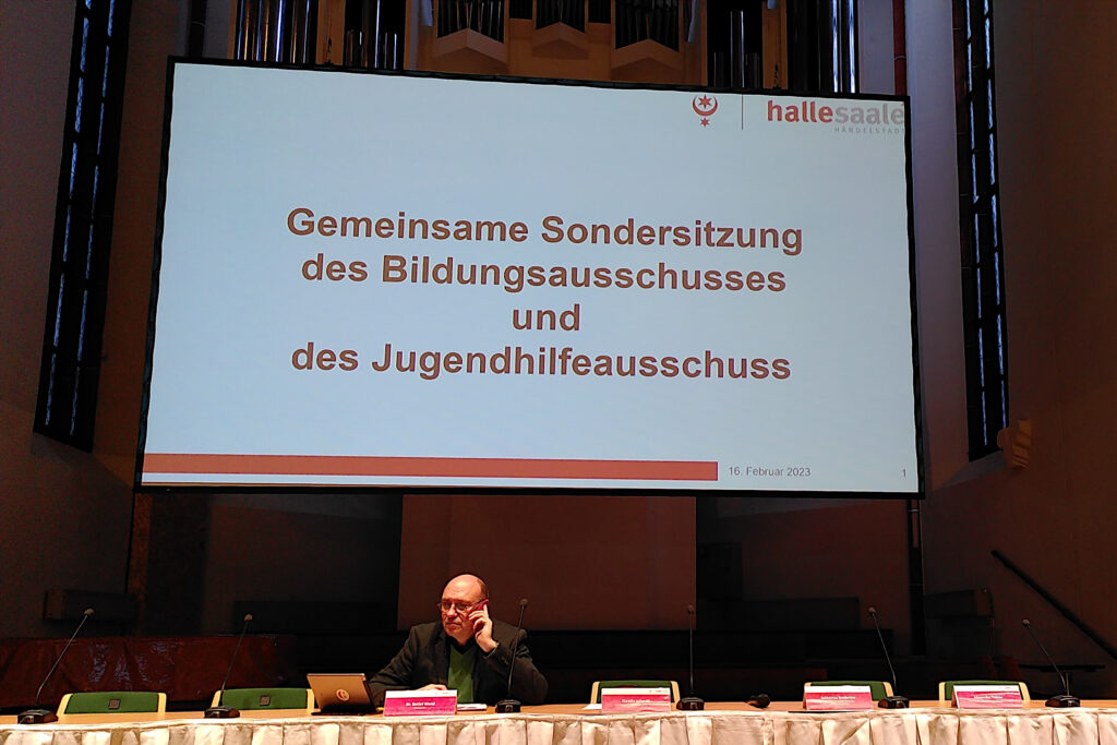 Stadtrat Detlef Wend auf dem Podium vor der Leinwand mit dem Schrifzug "Gemeinsame Sondersitzung des BIldungsausschusses und des Jugendhilfeausschusses"