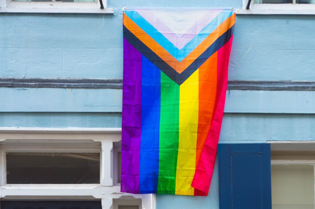 Eine vertikal an einer babyblauen Hausfassade hängende Pride Flagge in der Progress Version (mit Transfarben)