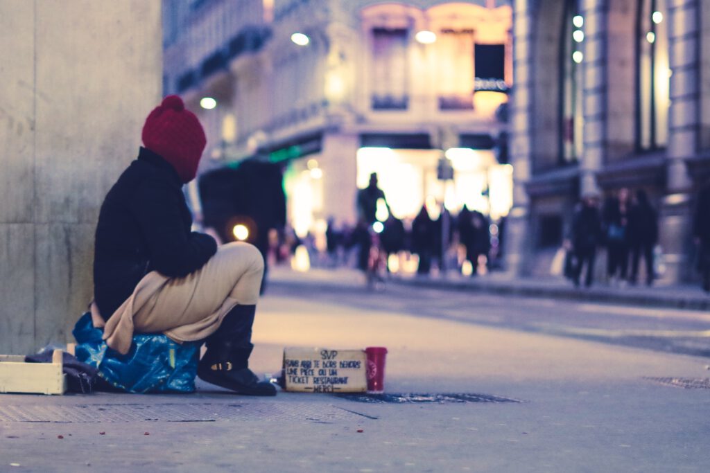 Seitlicher Blick auf eine wohnungslose Person mit roter Wintermütze, die in einer Fußgängerzone sitzt.