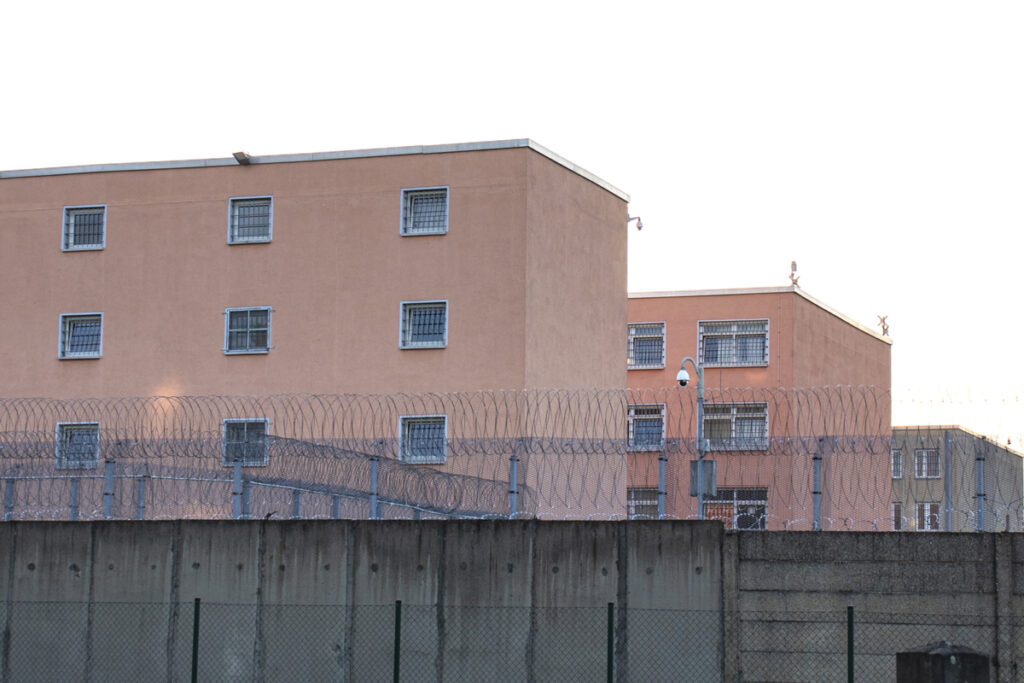 Blick auf die Zellenblöcke der JVA Frohe Zukunft über eine mit Stacheldraht besetzte Mauer hinweg